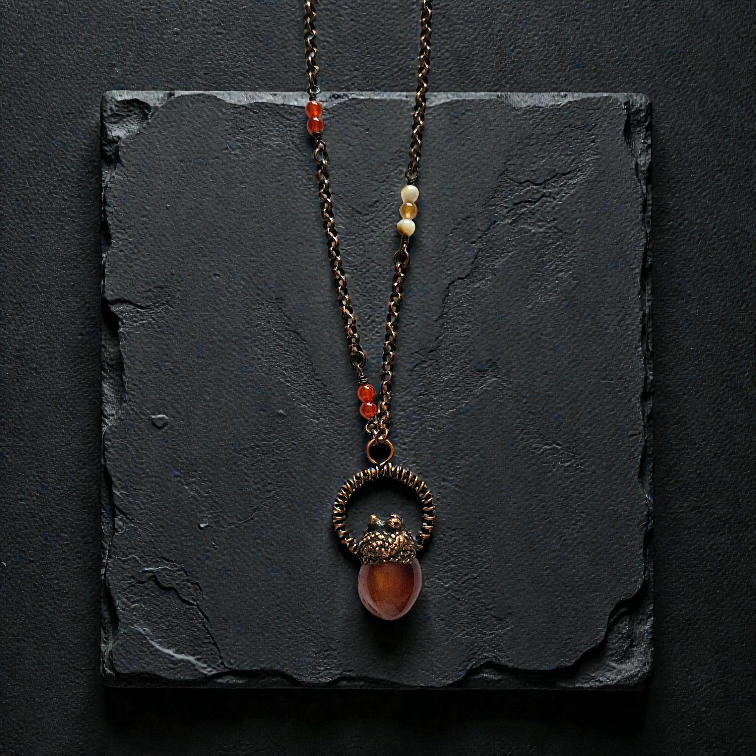 A copper electroformed carnelian pendant topped with an eternalized acorn cap and wire halo, displayed on a dark, moody textured background.