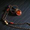 A copper electroformed carnelian pendant topped with an eternalized acorn cap and wire halo, displayed on a dark, moody textured background.