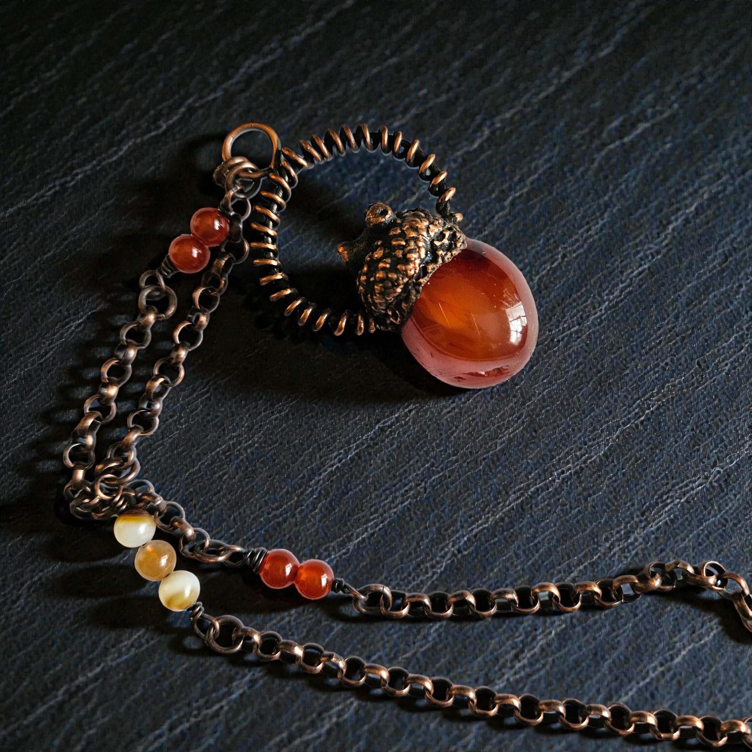 A copper electroformed carnelian pendant topped with an eternalized acorn cap and wire halo, displayed on a dark, moody textured background.
