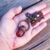 A hand holding the carnelian and copper acorn talisman to provide a size reference for the 2.5-inch pendant.