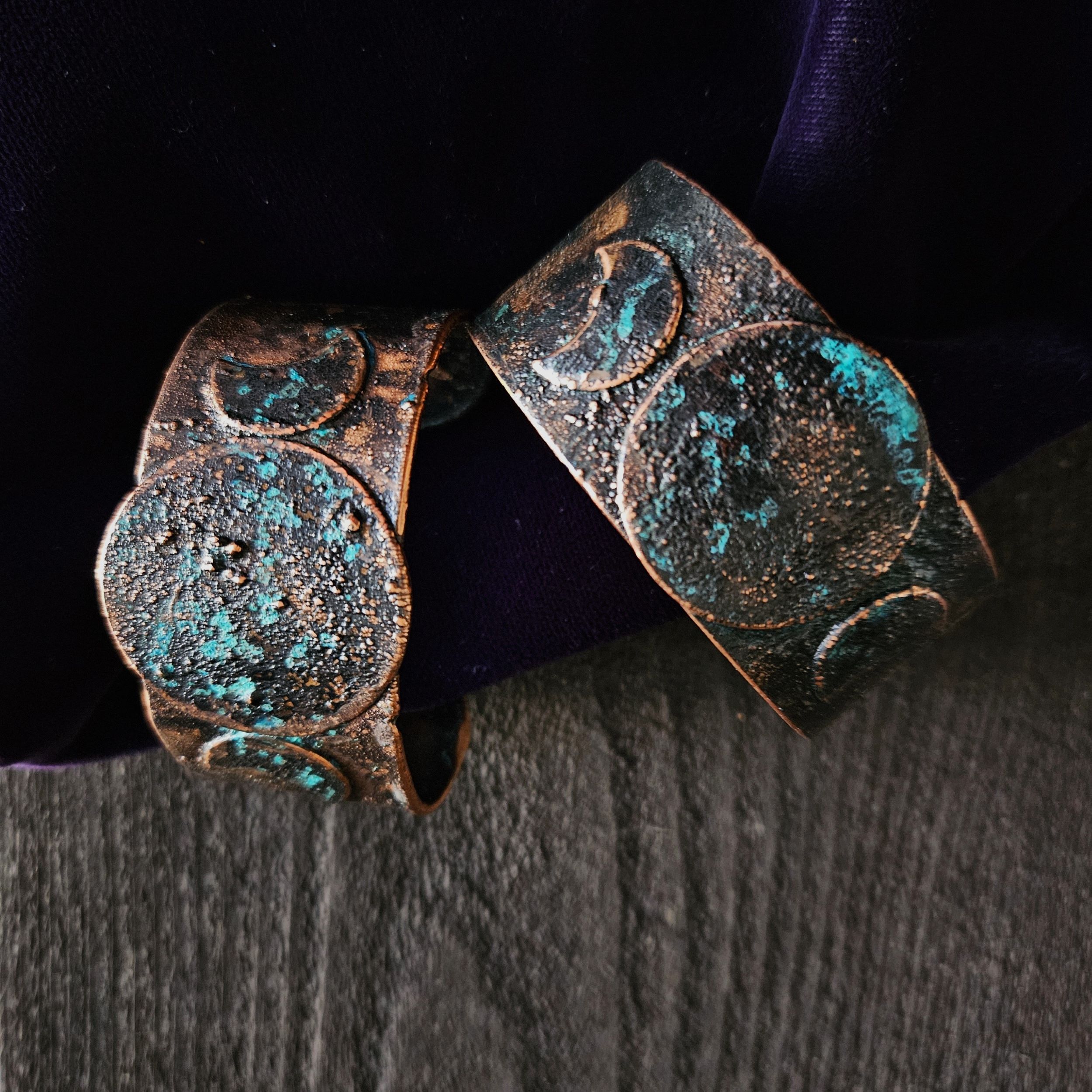 Hand-forged copper Triple Moon cuffs with a midnight blue patina on a background of dark wood and purple velvet.