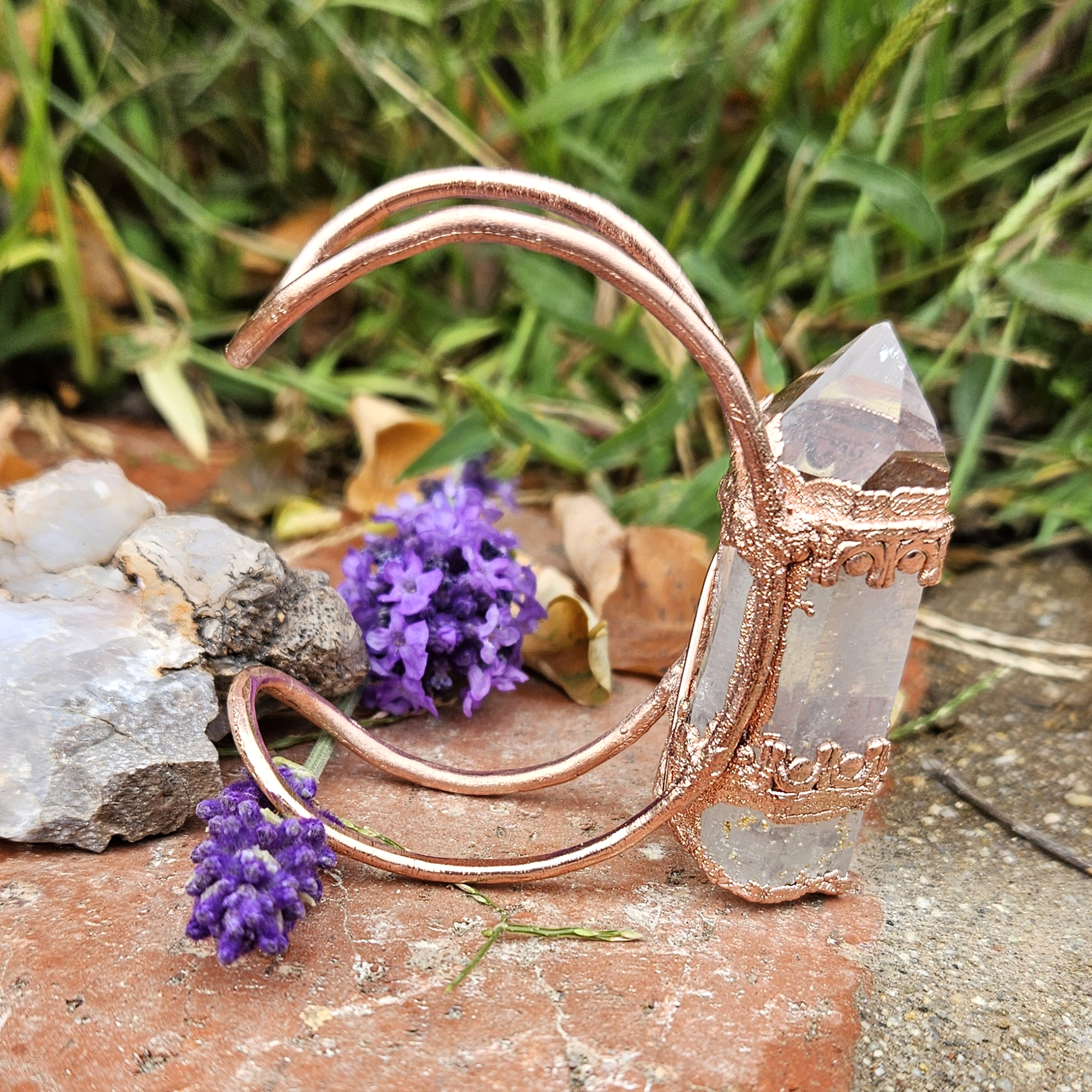 Hand-forged copper Archeron Cuff with a large vertical quartz point, displayed outdoors with lavender and stones.