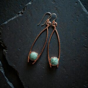 A pair of long copper electroformed earrings featuring raw blue-green aquamarine crystals on a dark, moody background.