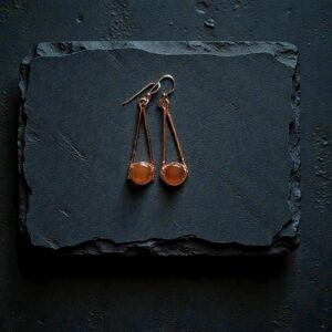 A pair of long copper electroformed earrings with orange carnelian stones resting on a dark, wet slate background.
