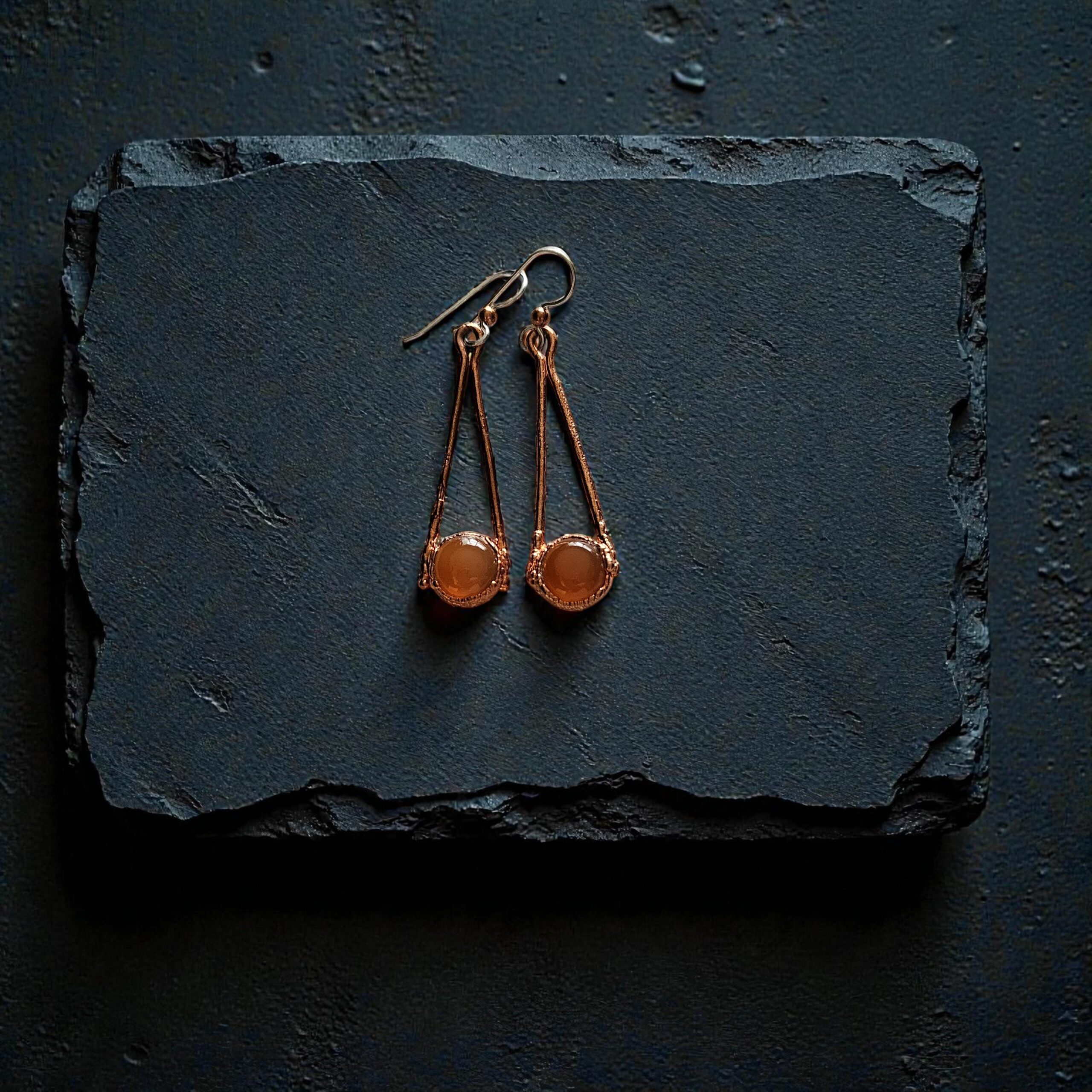 A pair of long copper electroformed earrings with orange carnelian stones resting on a dark, wet slate background.