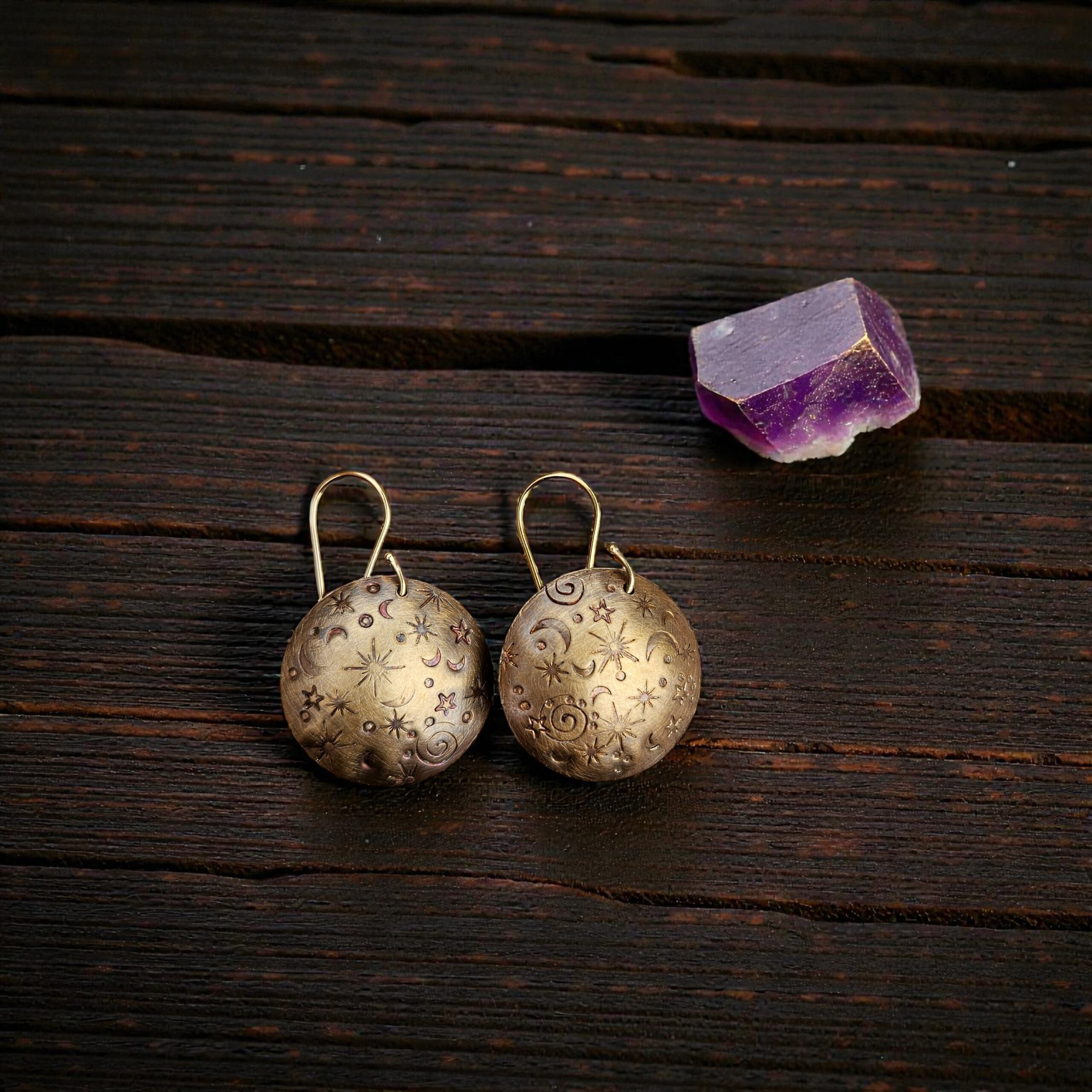 Hand-stamped Merlin's Gold brass earrings with an antiqued celestial moon and star pattern, resting on dark textured wood next to a purple amethyst crystal.
