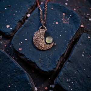 A dark-patina copper electroformed crescent moon amulet featuring a glowing green chalcedony stone.