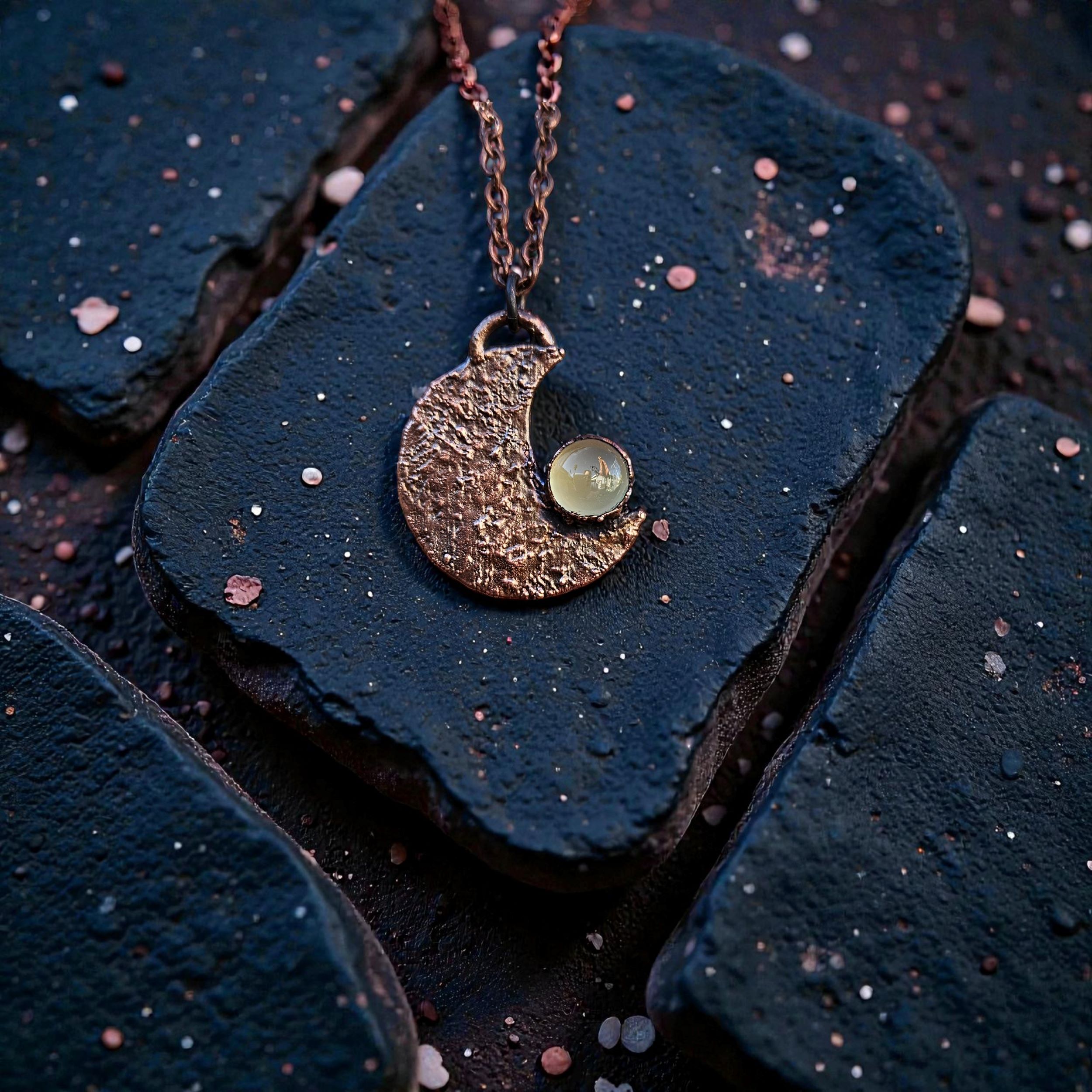 A dark-patina copper electroformed crescent moon amulet featuring a glowing green chalcedony stone.