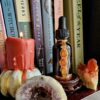 A 2oz amber glass dropper bottle encased in alchemical copper with three carnelian cabochons, styled on a bookshelf next to a red dripping candle and an amethyst geode.