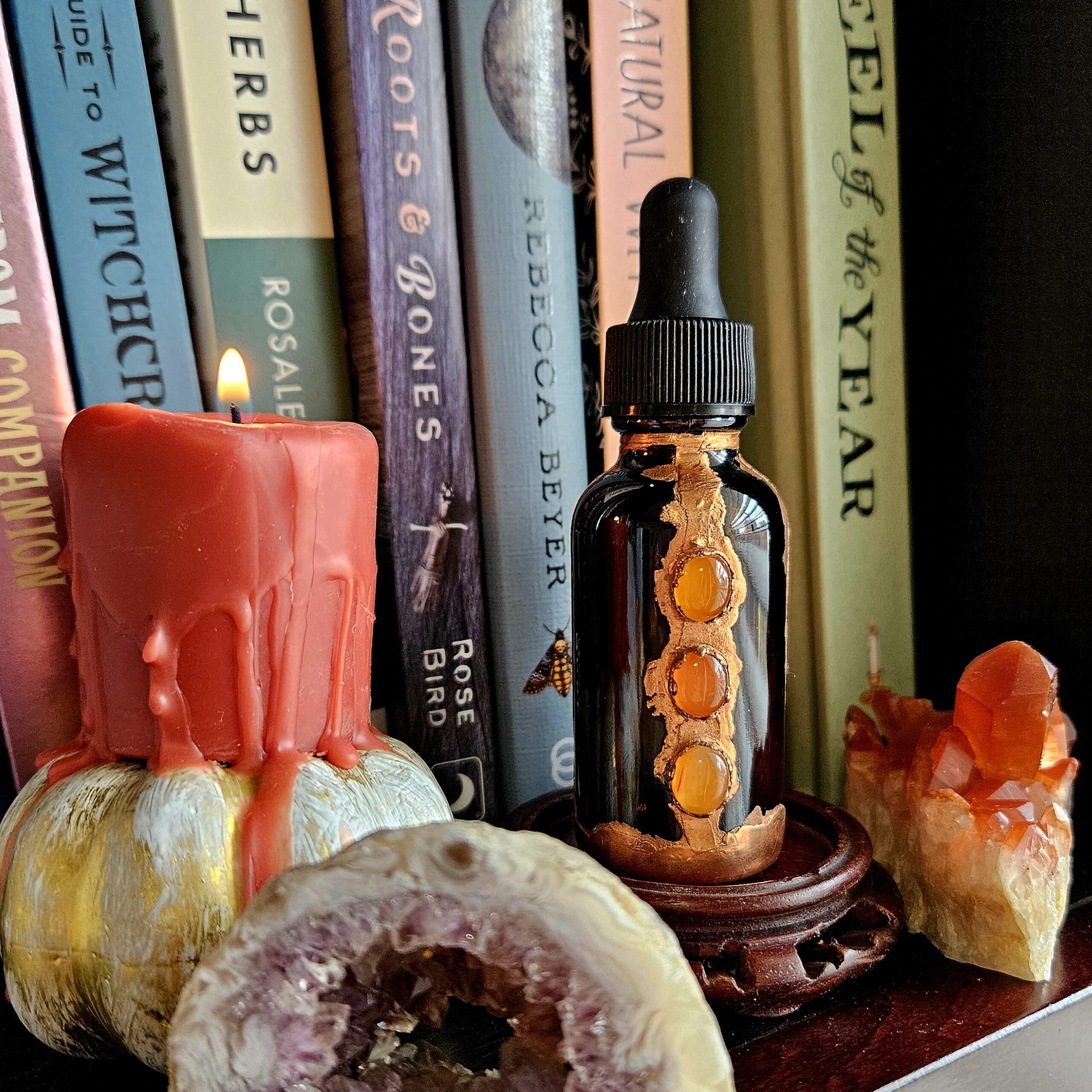 A 2oz amber glass dropper bottle encased in alchemical copper with three carnelian cabochons, styled on a bookshelf next to a red dripping candle and an amethyst geode.