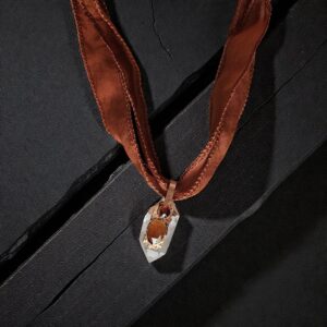 A handcrafted Solar Hearth Amulet featuring a clear quartz point topped with two orange hessonite garnets, encased in organic copper and suspended from a rust-colored silk ribbon on a dark, textured wood background.