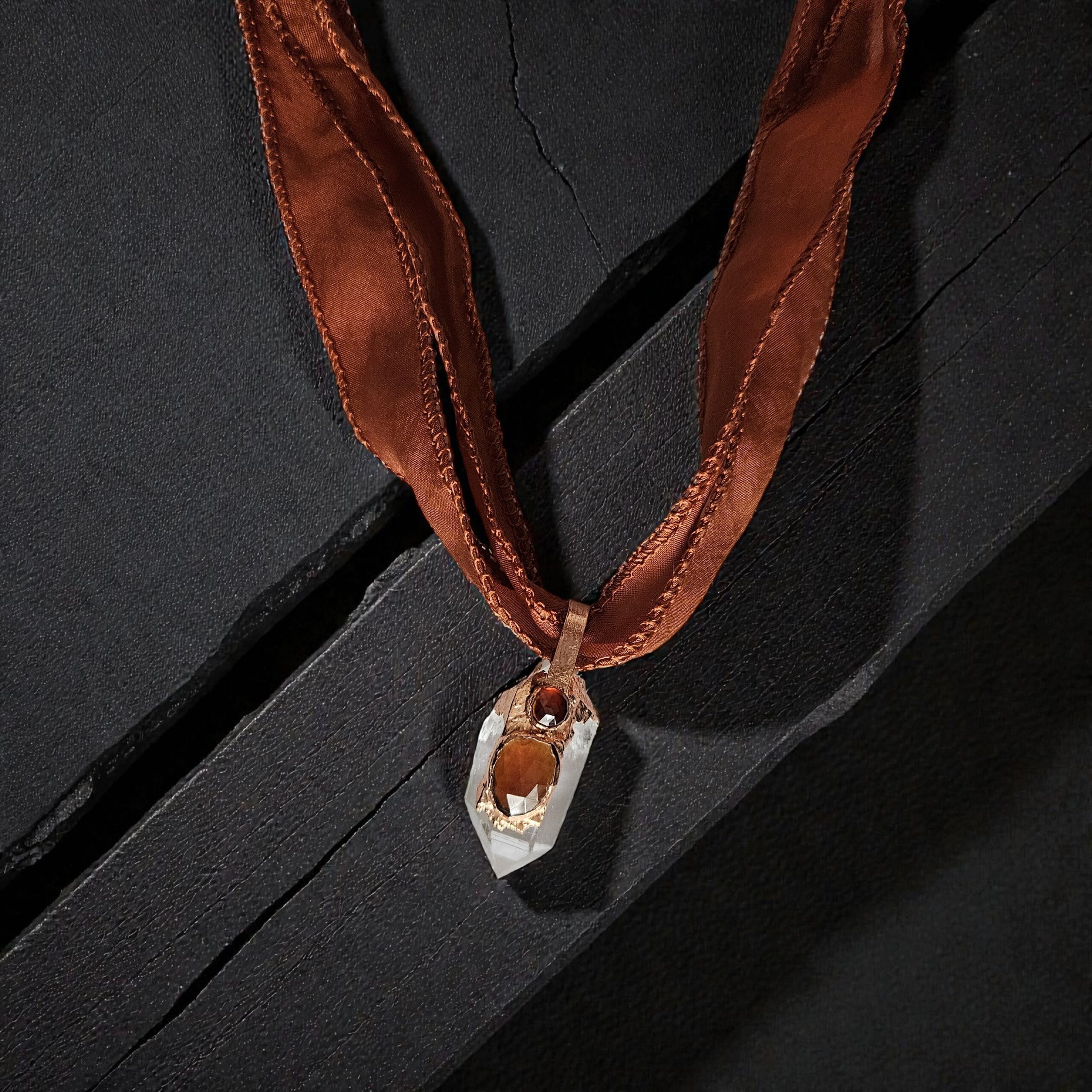 A handcrafted Solar Hearth Amulet featuring a clear quartz point topped with two orange hessonite garnets, encased in organic copper and suspended from a rust-colored silk ribbon on a dark, textured wood background.