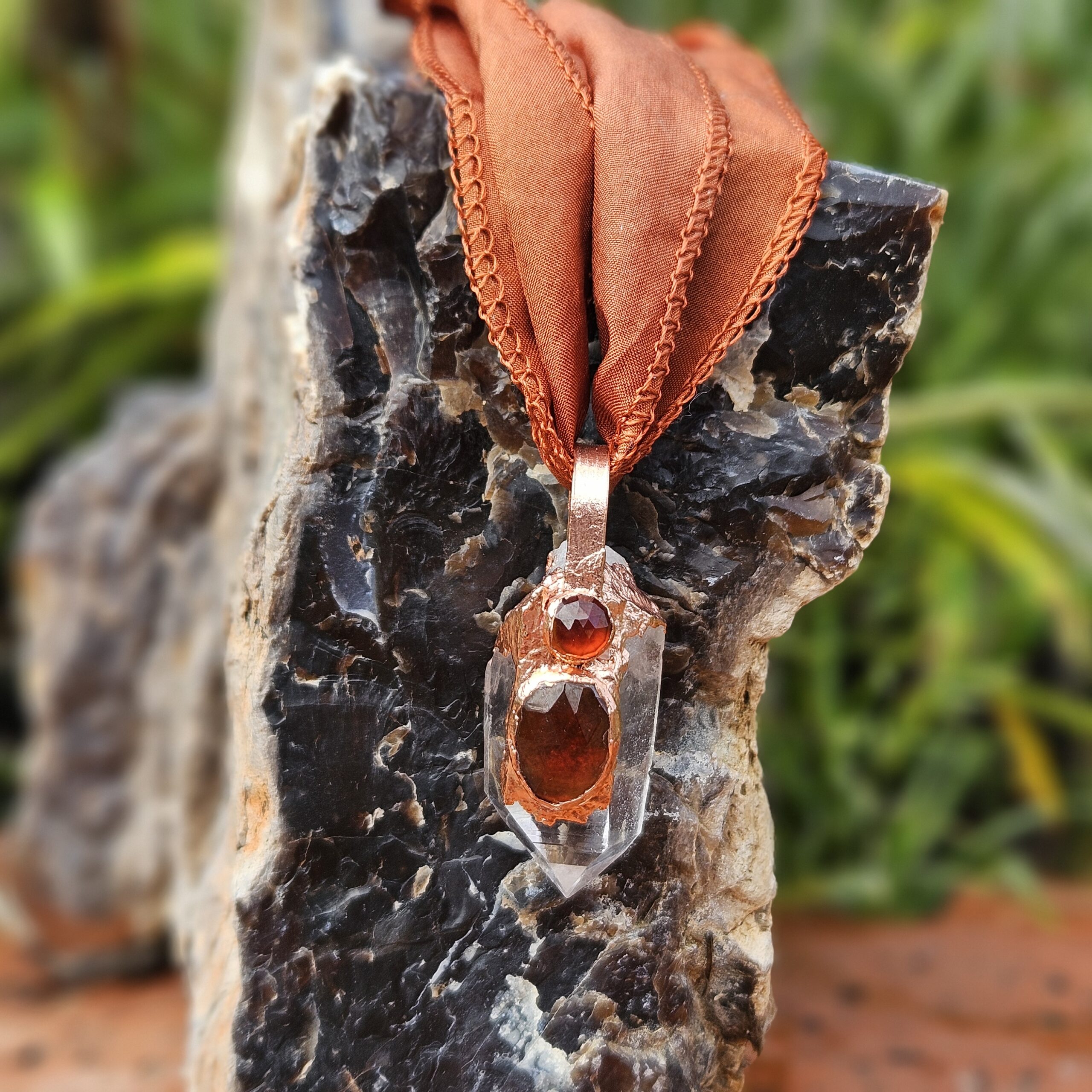 Close-up of a copper electroformed crystal amulet with a large faceted hessonite garnet and a smaller round garnet set into a clear quartz point.