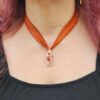 The Solar Hearth Amulet shown worn on a model, illustrating the length of the rust silk ribbon and the scale of the copper and quartz pendant against the neckline.