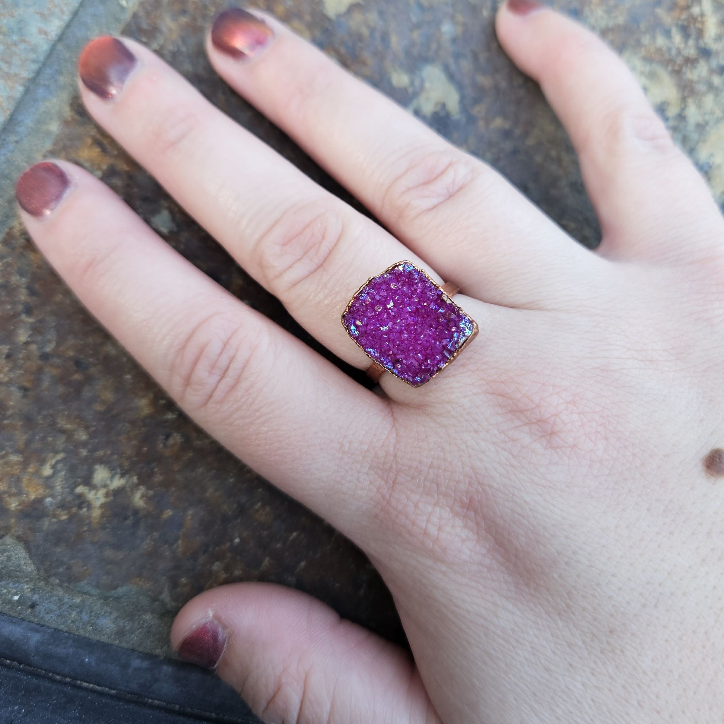 A close-up modeled shot of the Blushing Edge Druzy ring worn on a hand against a stone background. The ring features a large, rectangular magenta-pink druzy stone with a sparkling, crystalline surface and an electroformed copper edge. The vibrant pink of the stone is emphasized by natural outdoor lighting and coordinates with the wearer's shimmering reddish-brown manicure.