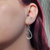 A close-up modeled shot of a Forged Compassion Talisman earring, featuring a hand-hammered sterling silver teardrop frame. A small, round, faceted pink gemstone is bezel-set at the bottom of the open frame, which hangs from a simple silver ear wire. The bright, reflective texture of the silver stands out against the wearer’s vibrant pink hair in a natural light setting.