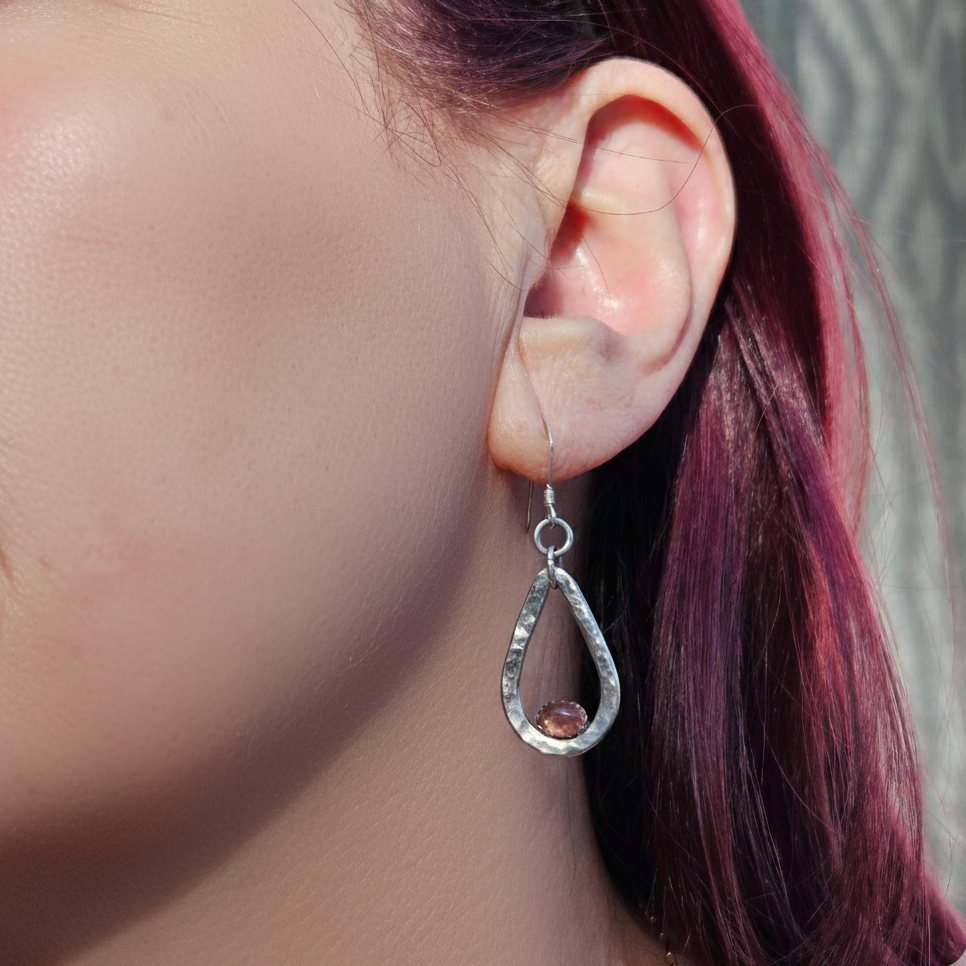 A close-up modeled shot of a Forged Compassion Talisman earring, featuring a hand-hammered sterling silver teardrop frame. A small, round, faceted pink gemstone is bezel-set at the bottom of the open frame, which hangs from a simple silver ear wire. The bright, reflective texture of the silver stands out against the wearer’s vibrant pink hair in a natural light setting.