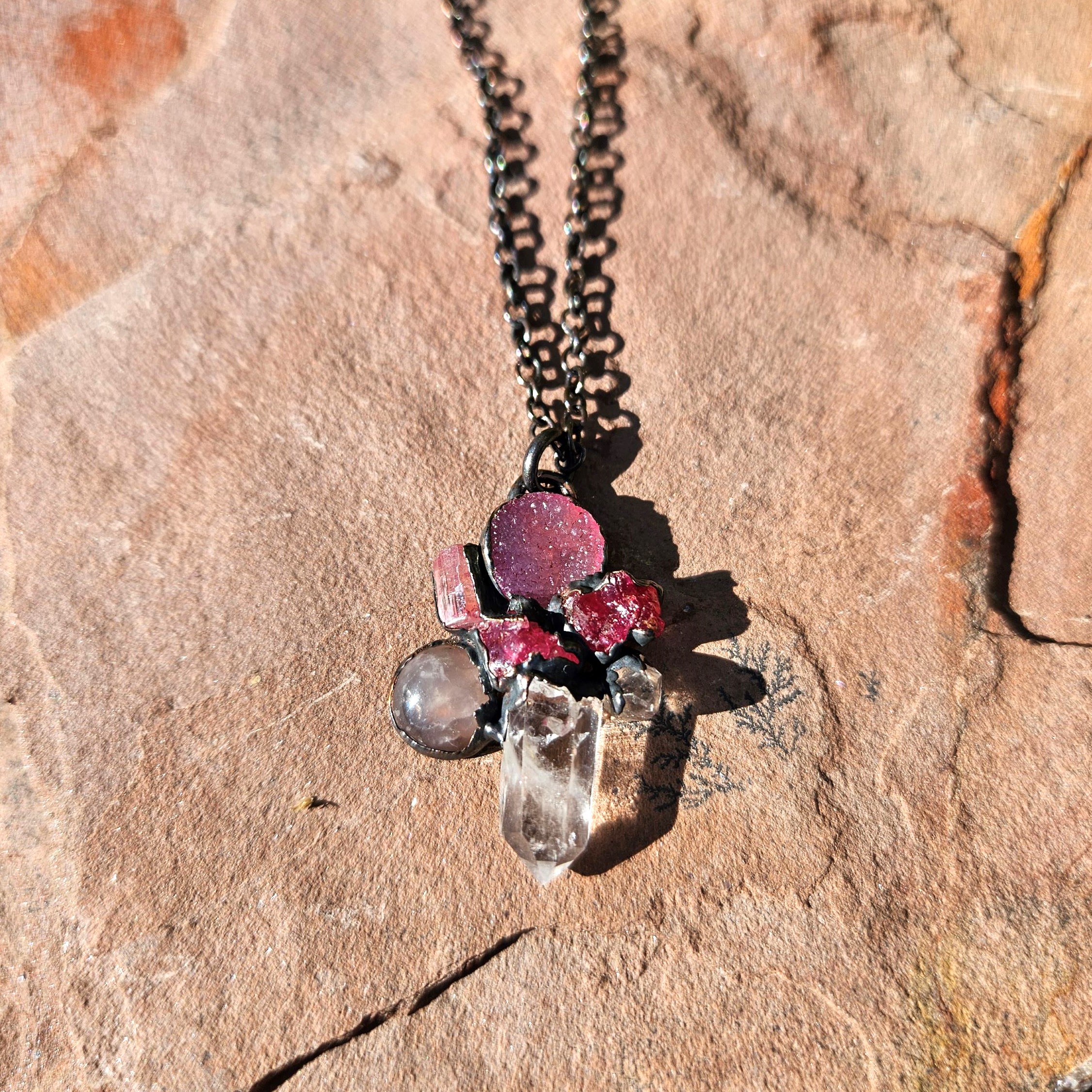 A moody, multi-stone pink talisman pendant featuring raw spinel and rose quartz, cast in deep shadows on a textured sandstone surface.