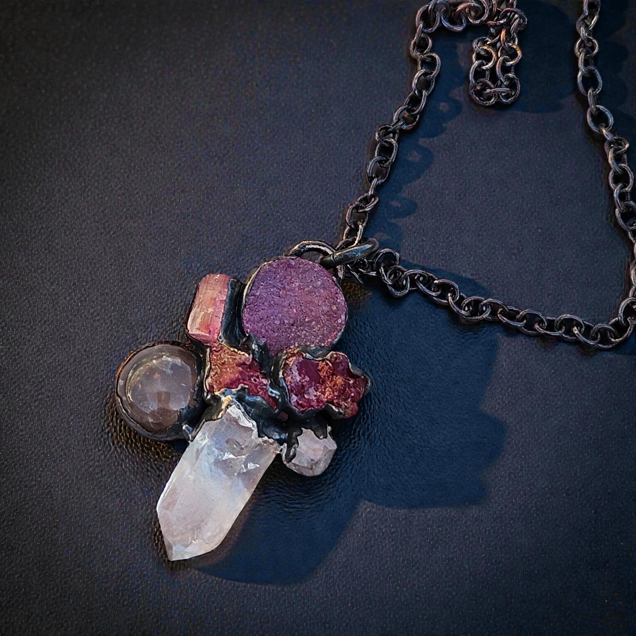 A moody, multi-stone pink talisman pendant featuring raw spinel and rose quartz, cast in deep shadows on a textured sandstone surface.