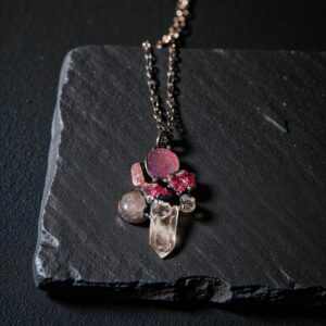 A dark and mysterious pink gemstone cluster pendant displayed on a black slate coaster, highlighting its ancient relic aesthetic.