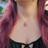 A modeled, close-up shot of the Pink Aura Diamond necklace, featuring a diamond-shaped pink aura druzy stone set in a minimalist gold-toned bezel. The necklace rests delicately at the collarbone, with the iridescent pink and purple facets of the druzy catching the warm, natural sunlight. The vibrant pink of the stone is complemented by the wearer’s coordinating pink hair in the background.