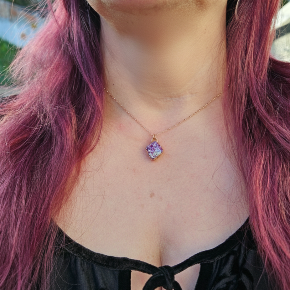 A modeled, close-up shot of the Pink Aura Diamond necklace, featuring a diamond-shaped pink aura druzy stone set in a minimalist gold-toned bezel. The necklace rests delicately at the collarbone, with the iridescent pink and purple facets of the druzy catching the warm, natural sunlight. The vibrant pink of the stone is complemented by the wearer’s coordinating pink hair in the background.