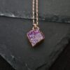 A diamond-shaped pink aura druzy necklace with iridescent crystalline sparkle displayed on a textured black stone surface.