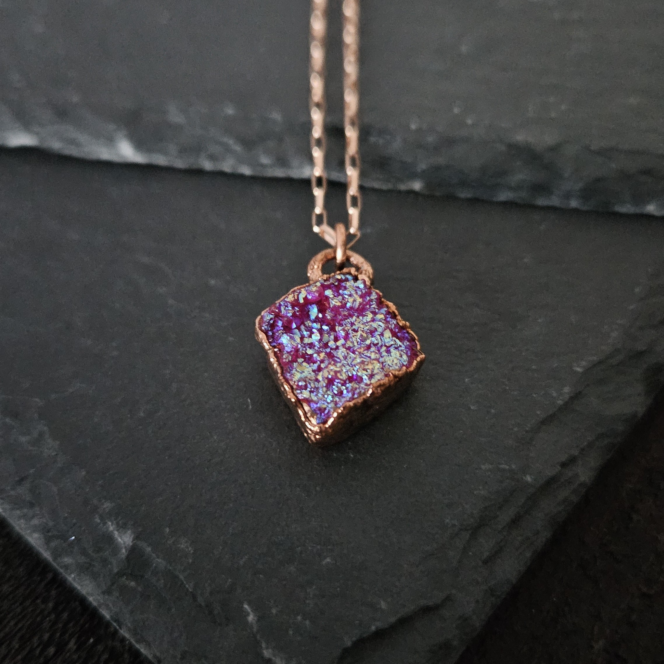 A diamond-shaped pink aura druzy necklace with iridescent crystalline sparkle displayed on a textured black stone surface.