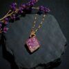 Pink aura druzy diamond pendant styled on a dark slate fragment with a sprig of purple flowers, capturing a moody ritual aesthetic.