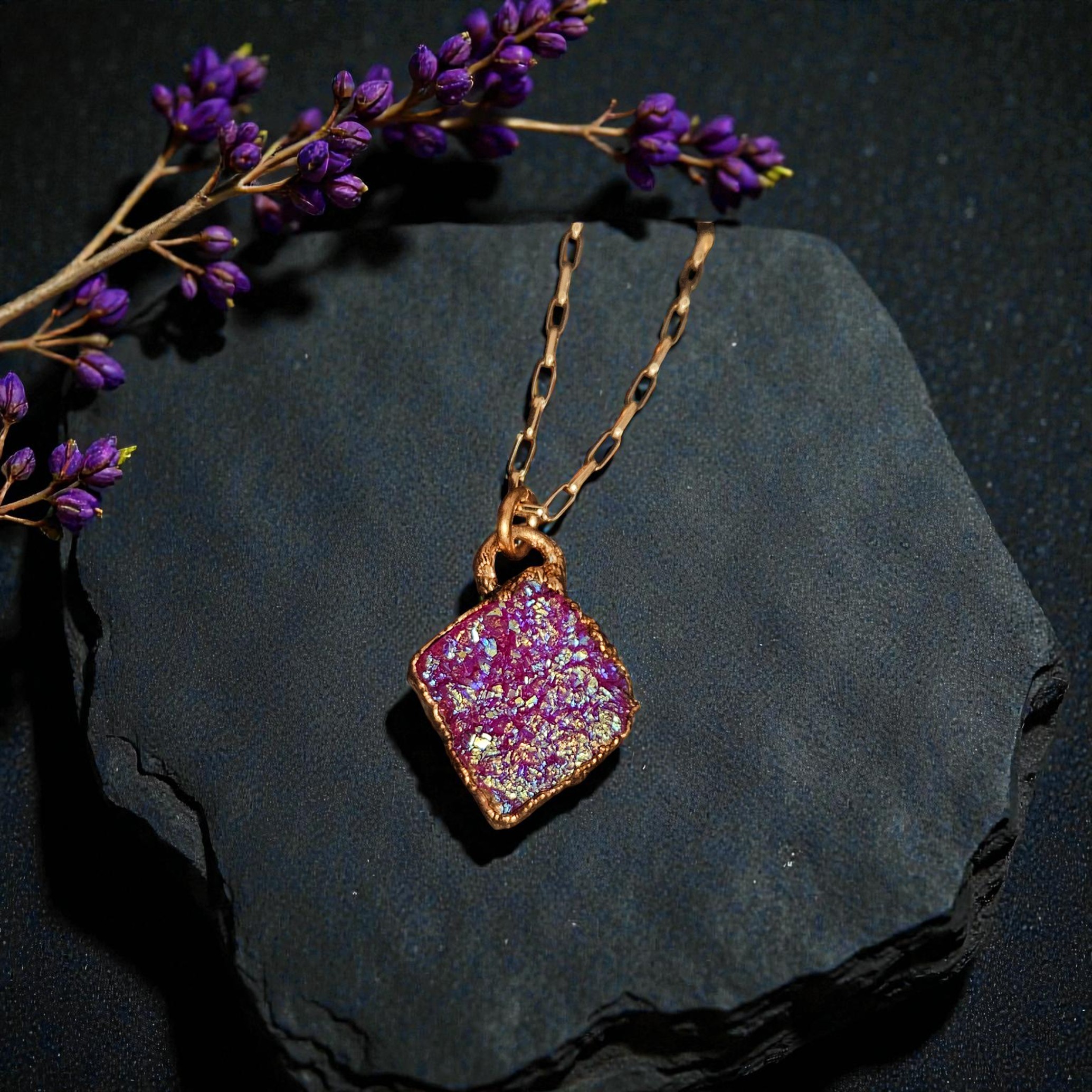 Pink aura druzy diamond pendant styled on a dark slate fragment with a sprig of purple flowers, capturing a moody ritual aesthetic.