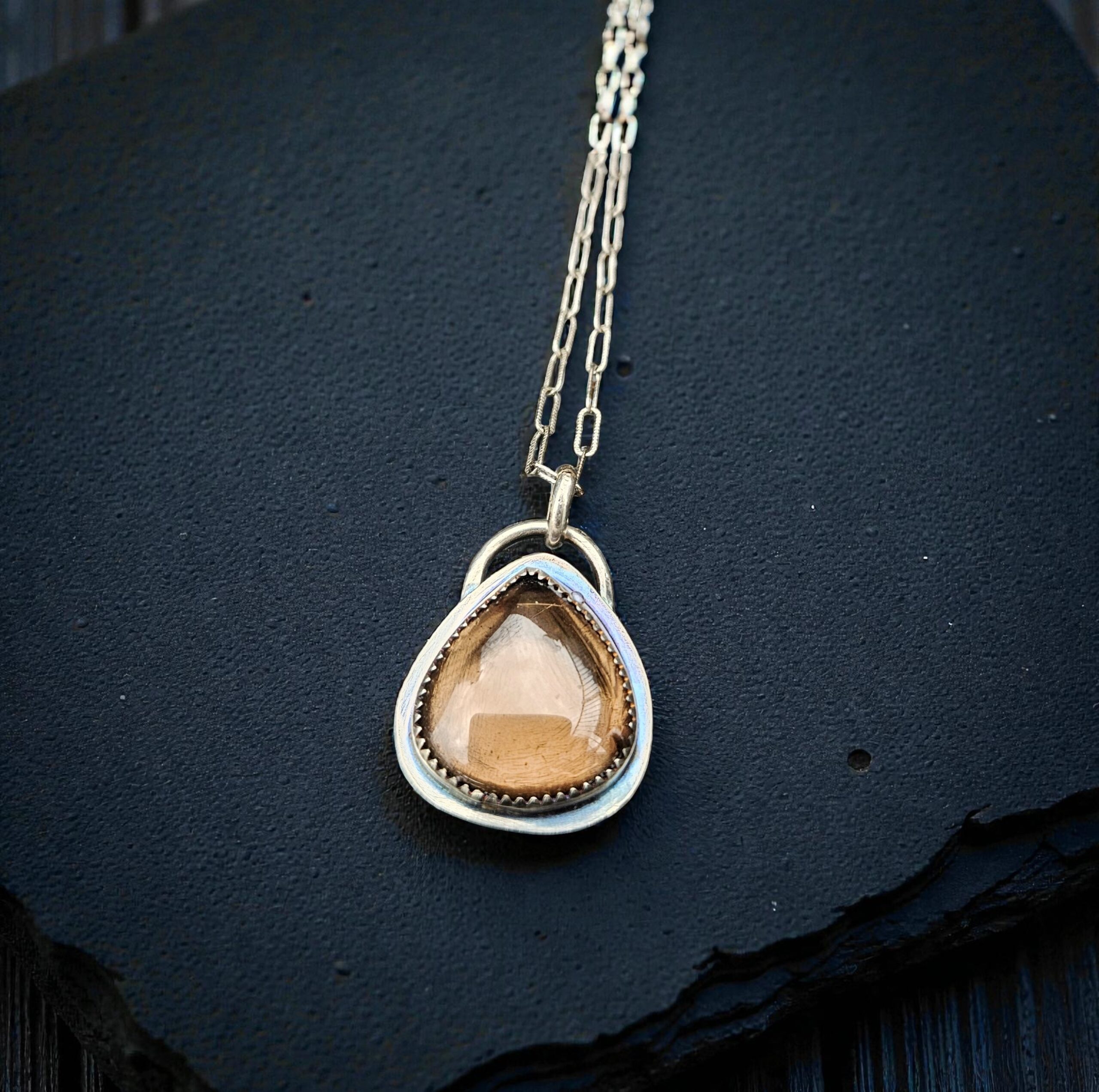 A teardrop-shaped smoky quartz cabochon set in a sterling silver saw-tooth bezel, worn as a pendant on a delicate silver chain.