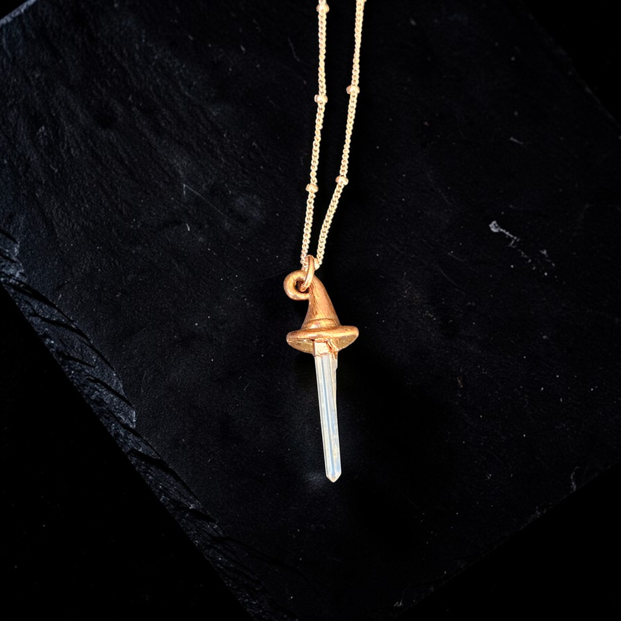 Handcrafted copper electroformed witch hat necklace with a long, clear quartz crystal point as the peak. The pendant is shown against a dark slate background, attached to a rose gold filled chain.