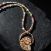 Flat lay of a hand-grown copper electroformed ammonite fossil pendant featuring a flash of blue labradorite at its center and a raw quartz crystal point at the base. The pendant hangs from an asymmetrical, multicolored strand of natural gemstone beads against a dark slate background.
