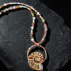 Flat lay of a hand-grown copper electroformed ammonite fossil pendant featuring a flash of blue labradorite at its center and a raw quartz crystal point at the base. The pendant hangs from an asymmetrical, multicolored strand of natural gemstone beads against a dark slate background.