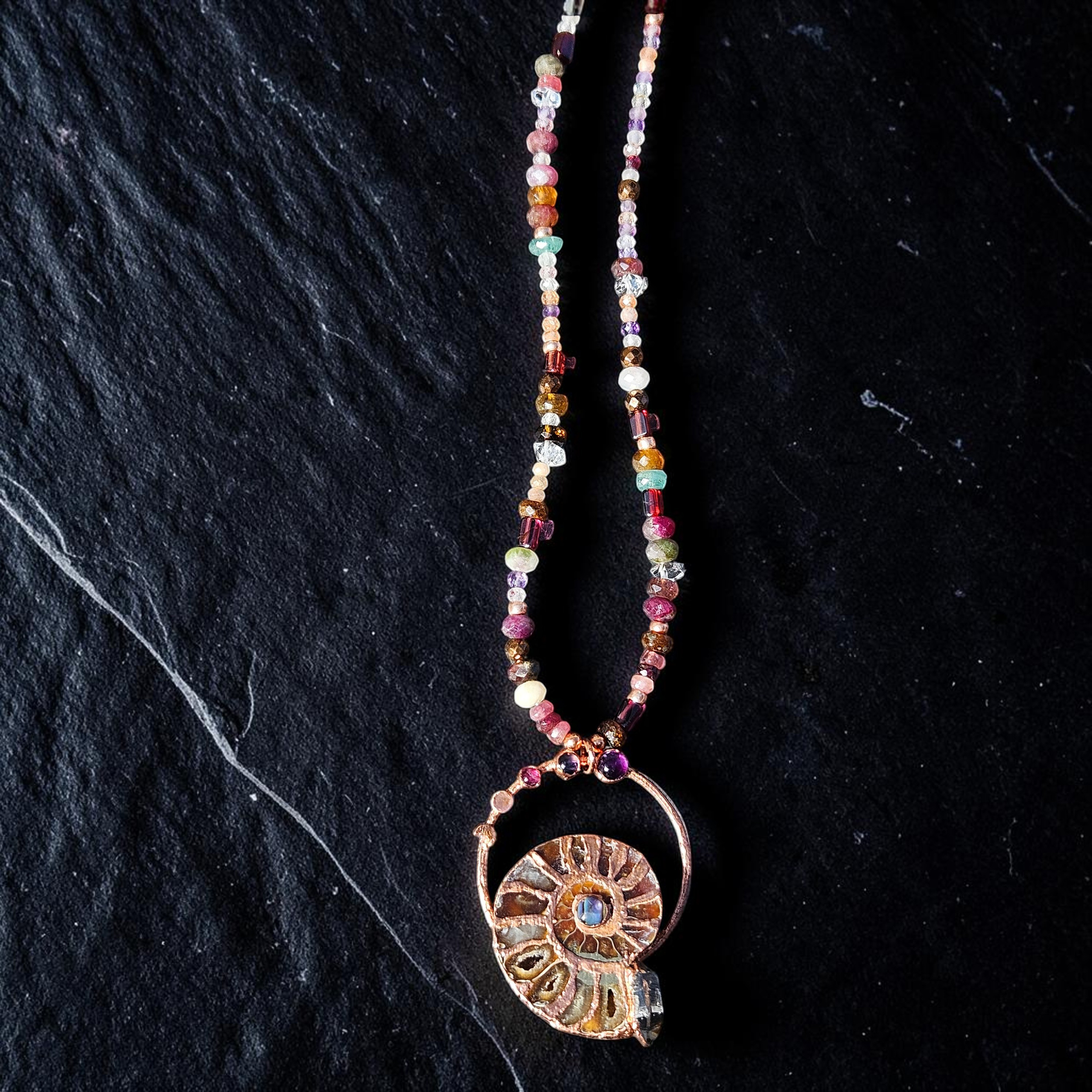 High-contrast overhead view highlighting the intricate, textured copper metalwork framing the natural chambers of the cut ammonite fossil and its vibrant multi-gemstone accent strand.