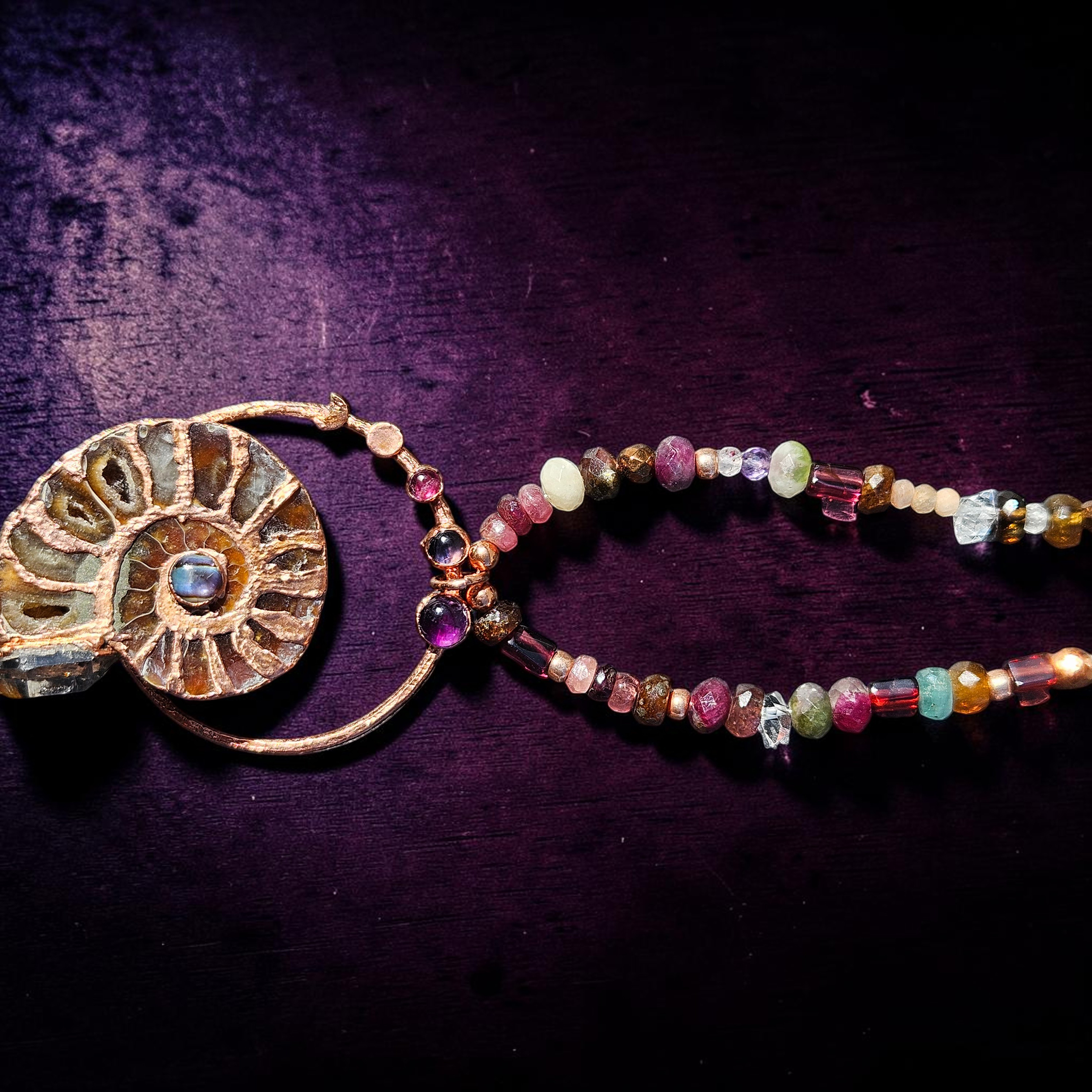 Macro close-up shot showcasing the fine details of the copper electroformed setting, highlighting the small garnet, iolite, and amethyst cabochons embedded at the neck loop, alongside the rich textures of the natural beaded strand.
