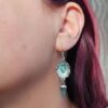 A close-up profile view of a model wearing the shorter apatite side of the asymmetrical earrings, showing the 1.75-inch drop length, sterling silver earwire, and the vibrant teal-blue crystal drop.