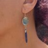 A close-up profile view of a model wearing the longer blue kyanite side of the pair, showing the elegant, dramatic 2.125-inch drop length and the slender, deep blue crystalline blade.