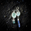 A flat-lay photograph on shimmering black sand showing a pair of asymmetrical dangle earrings. Each earring features a silver-framed limpet shell showing its smooth, pale sea-foam green interior. One suspends a short, chunky raw neon blue apatite crystal, and the other suspends a long, raw blue kyanite blade.
