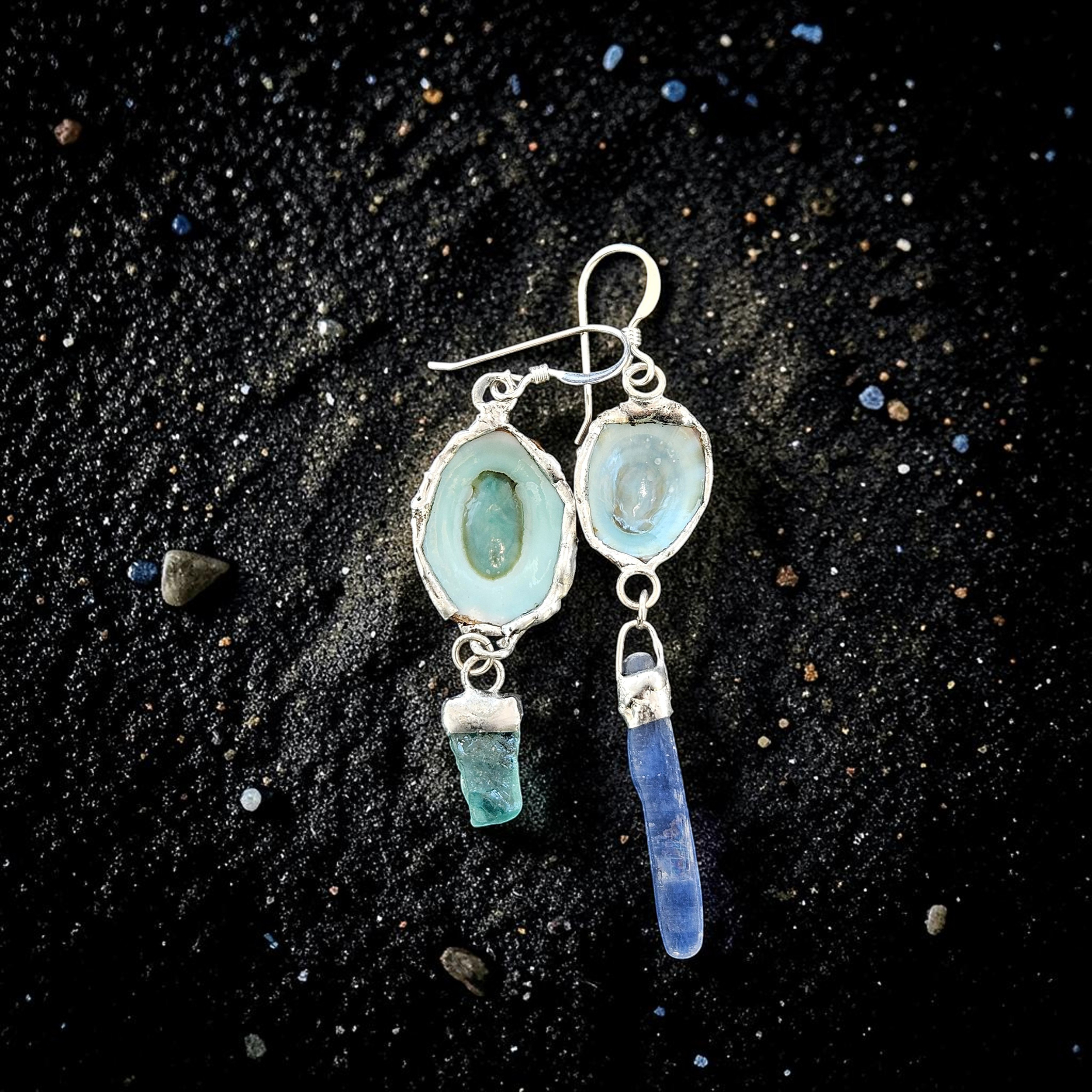 A flat-lay photograph on shimmering black sand showing a pair of asymmetrical dangle earrings. Each earring features a silver-framed limpet shell showing its smooth, pale sea-foam green interior. One suspends a short, chunky raw neon blue apatite crystal, and the other suspends a long, raw blue kyanite blade.
