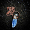 Overhead view of the blue kyanite talisman resting on shimmering black sand, showing the full length of the 18-inch bright copper link chain coiled neatly above the pendant.
