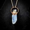 A flat lay product shot on a textured black slate background showing a long, raw blue kyanite crystal blade. The crystal is crowned with a round, carved mother-of-pearl moon face doublet under glass, securely encased in a highly textured copper electroformed orbit.