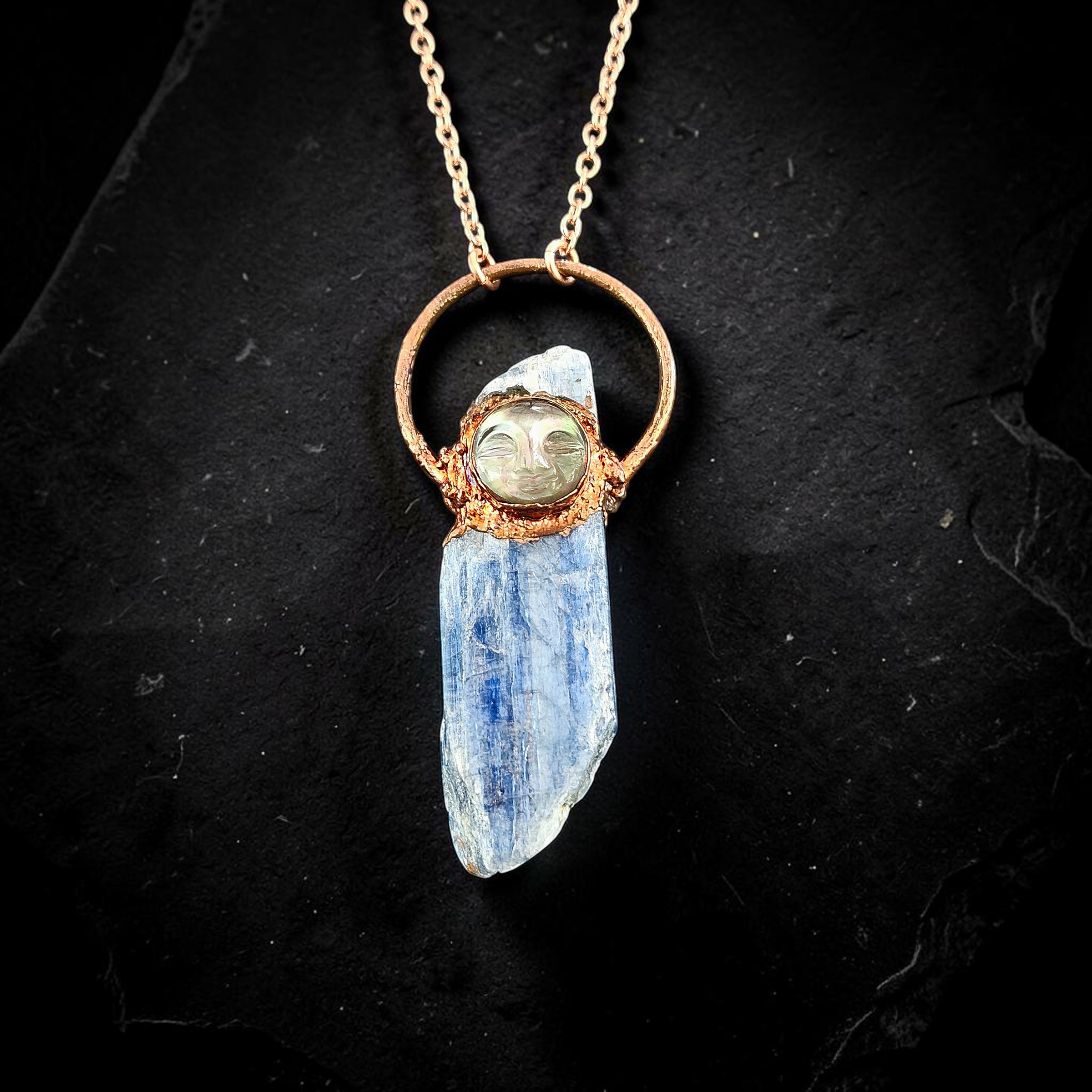 A flat lay product shot on a textured black slate background showing a long, raw blue kyanite crystal blade. The crystal is crowned with a round, carved mother-of-pearl moon face doublet under glass, securely encased in a highly textured copper electroformed orbit.