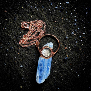 Overhead view of the blue kyanite talisman resting on shimmering black sand, showing the full length of the 18-inch bright copper link chain coiled neatly above the pendant.