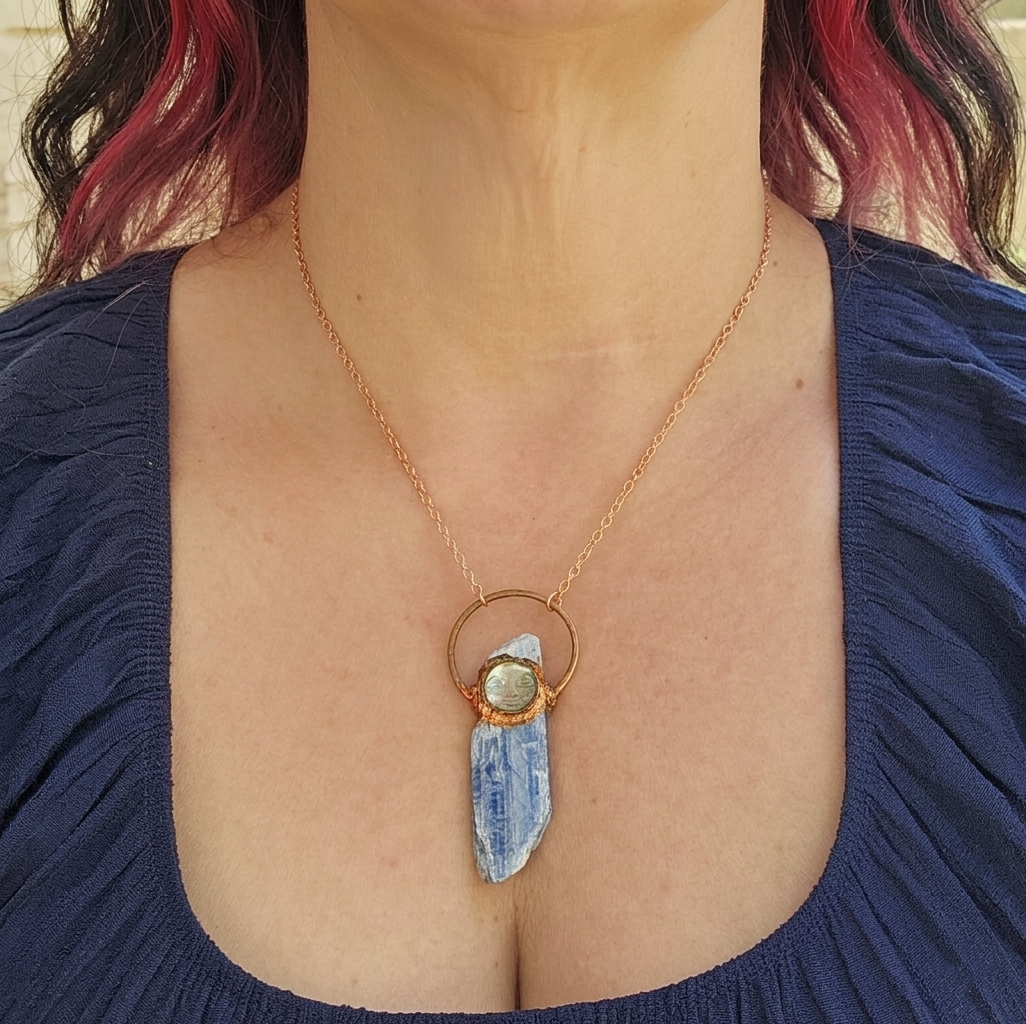 A modeled shot demonstrating the 2.75-inch vertical drape of the blue kyanite and carved moon face pendant, resting elegantly below the collarbone against a navy blue neckline.