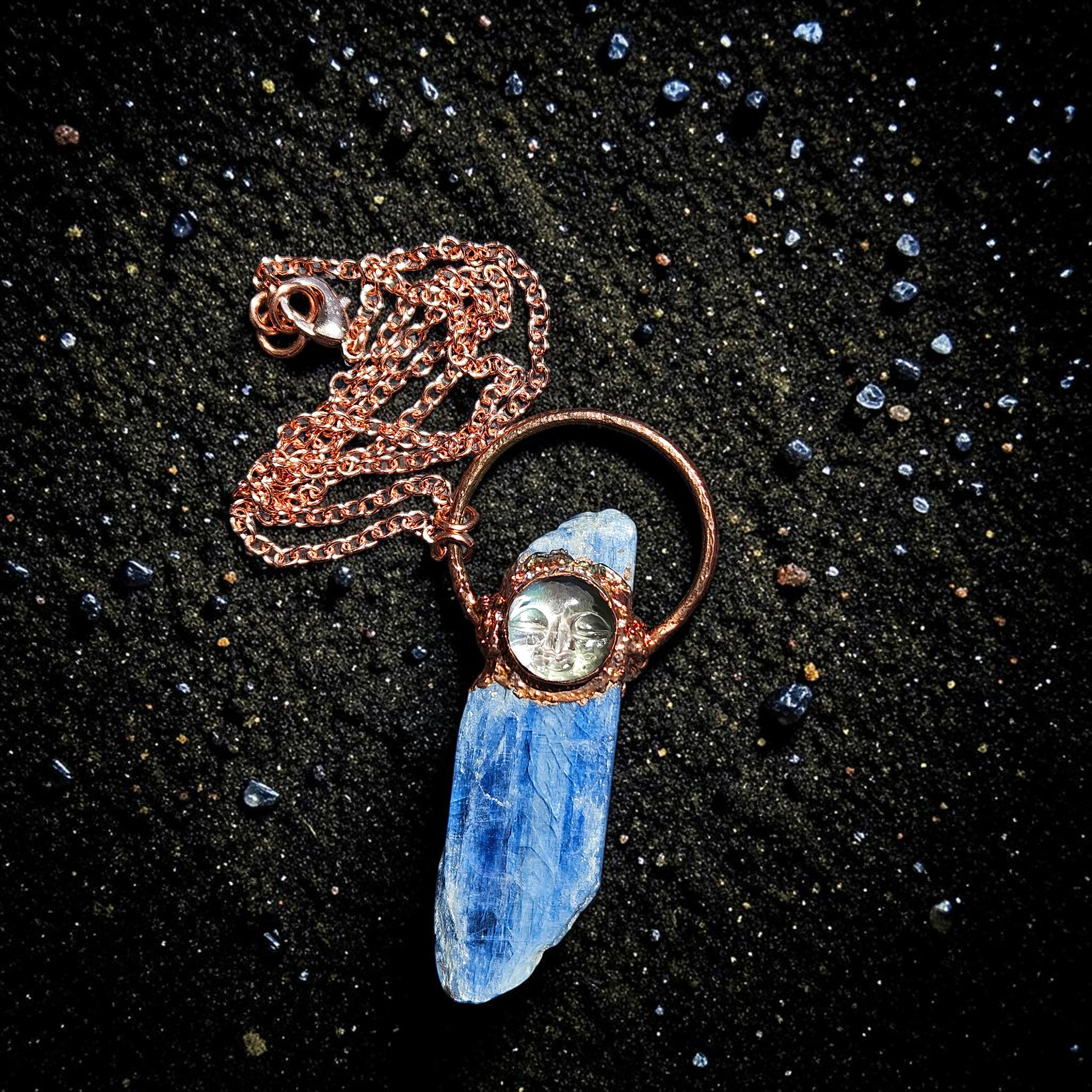 Overhead view of the blue kyanite talisman resting on shimmering black sand, showing the full length of the 18-inch bright copper link chain coiled neatly above the pendant.