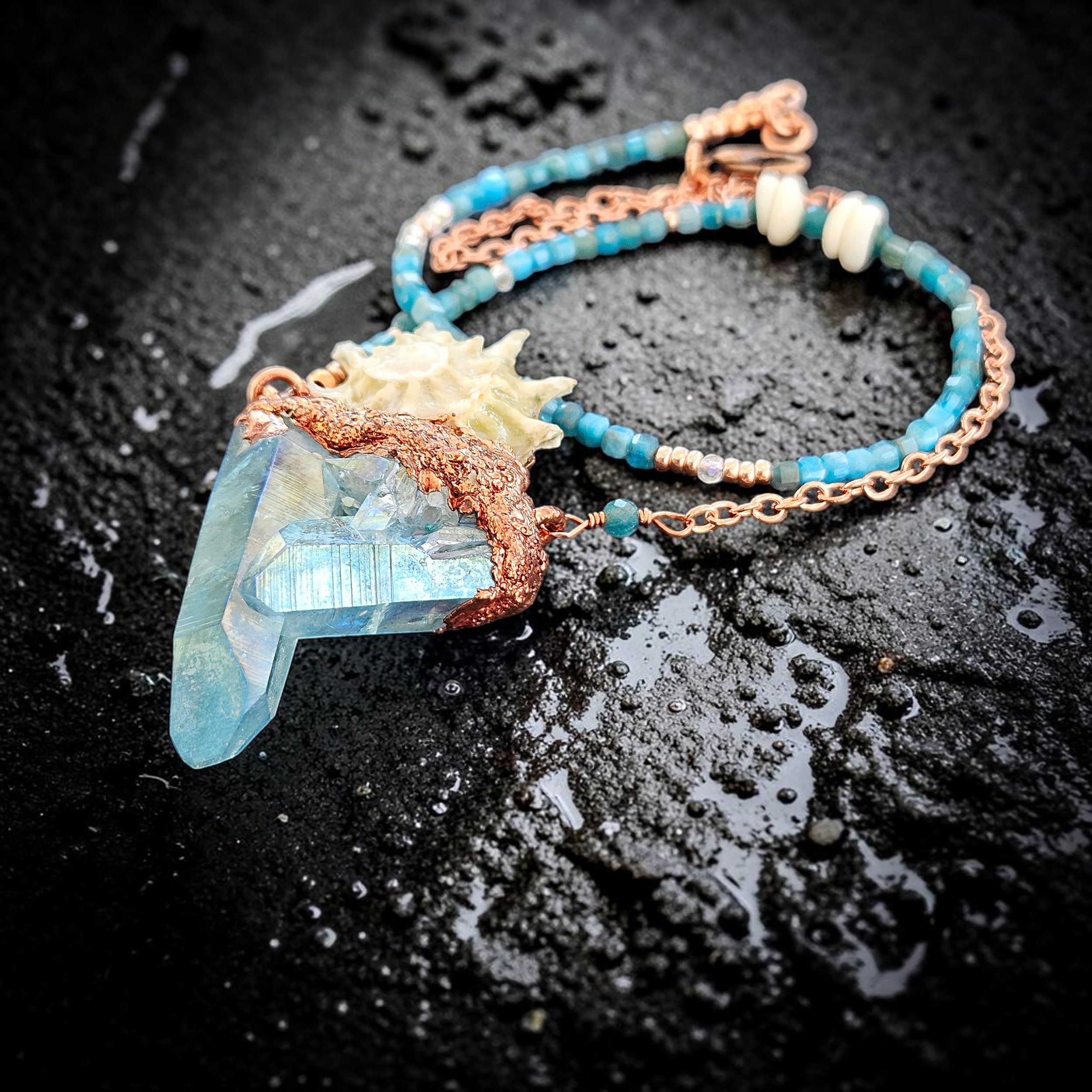 Flat lay shot on a dark slate background showing a vibrant, electric blue aqua aura crystal cluster pendant topped with a spiked Spiny Star Astraea shell. The necklace features a highly unique asymmetrical chain made of natural blue apatite cubes, white vintage puka shells, and copper link details.