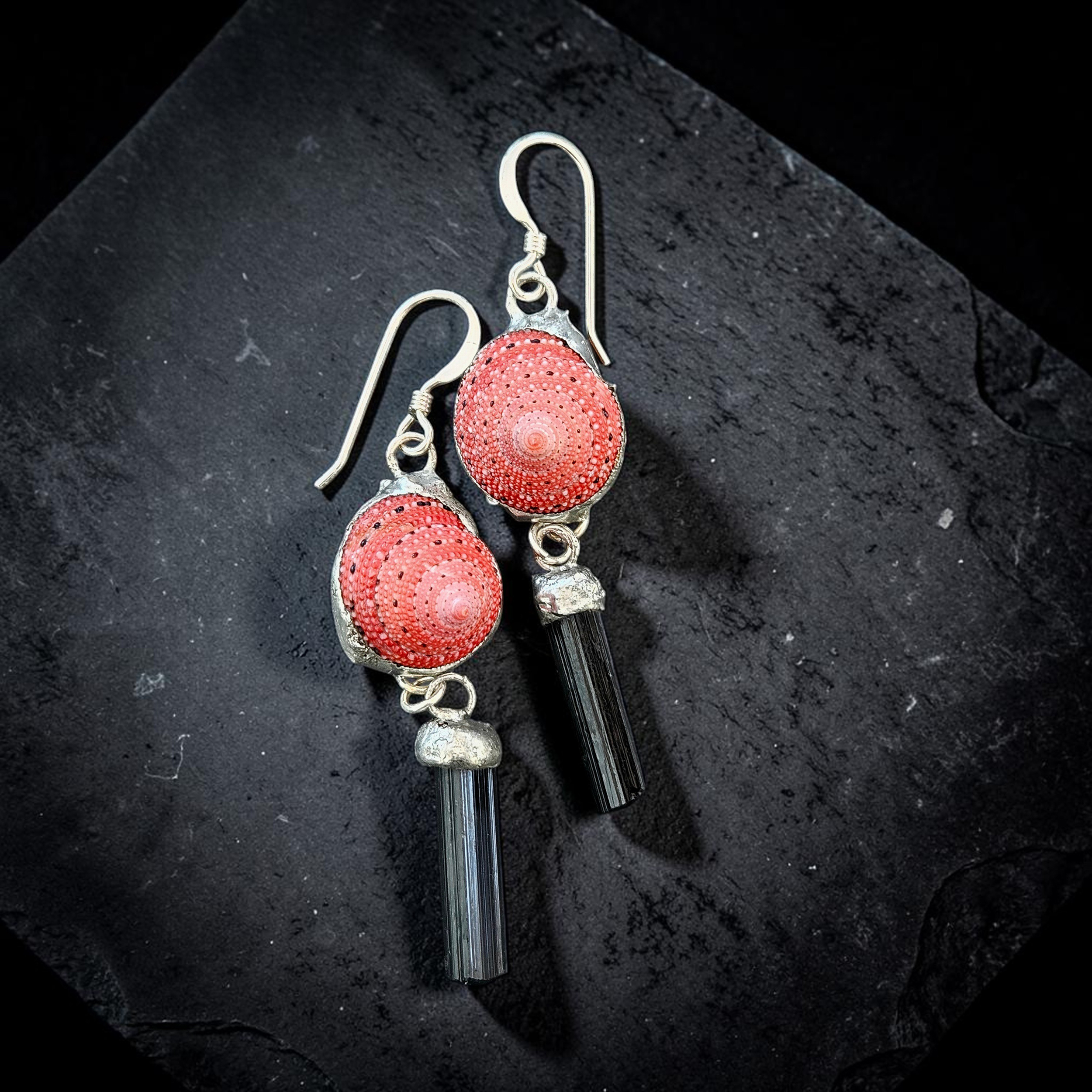 A flat-lay photograph on a dark slate background showing a pair of handmade dangle earrings. Each earring features a bright, coral-pink strawberry top shell with a dotted texture, set in silver metal work, with a raw, striated black tourmaline crystal rod dangling underneath.