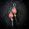 An overhead shot of the strawberry shell and black tourmaline earrings resting on glistening black volcanic sand, highlighting the silver metal texture and the rich color contrast of the pink shells and black stones.