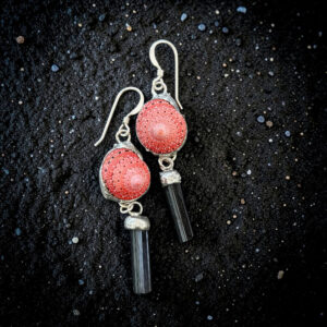 An overhead shot of the strawberry shell and black tourmaline earrings resting on glistening black volcanic sand, highlighting the silver metal texture and the rich color contrast of the pink shells and black stones.